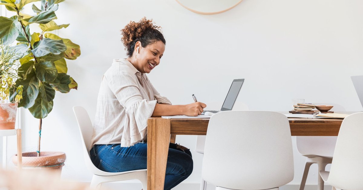 A smiling person sitting at their dining table and writing in a notebook next to an open laptop.