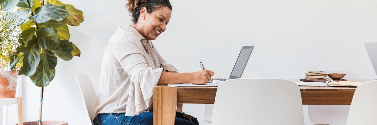 A smiling person sitting at their dining table and writing in a notebook next to an open laptop.