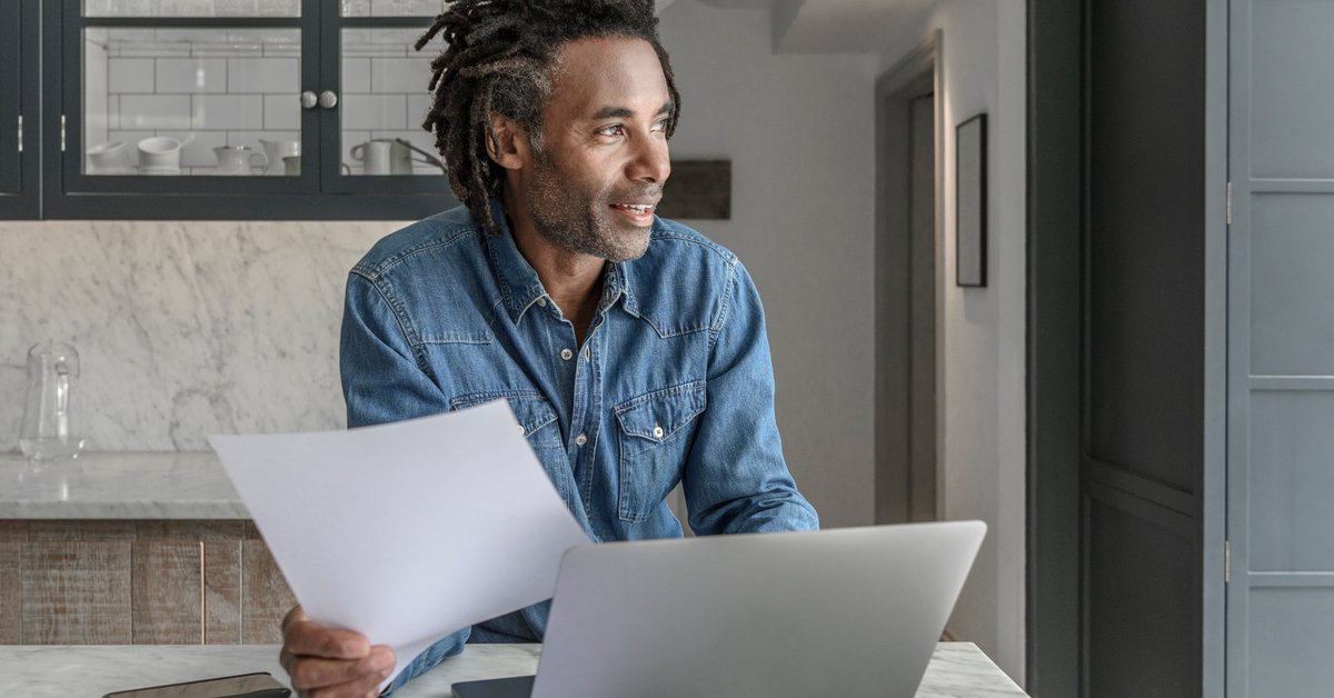 A person looking at paperwork and a laptop while standing at the kitchen counter.