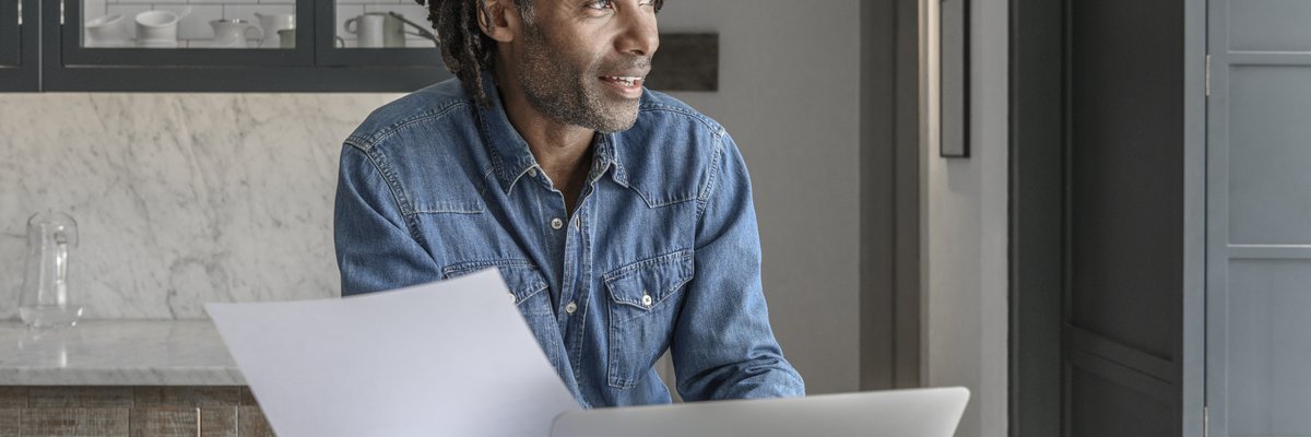 A person looking at paperwork and a laptop while standing at the kitchen counter.