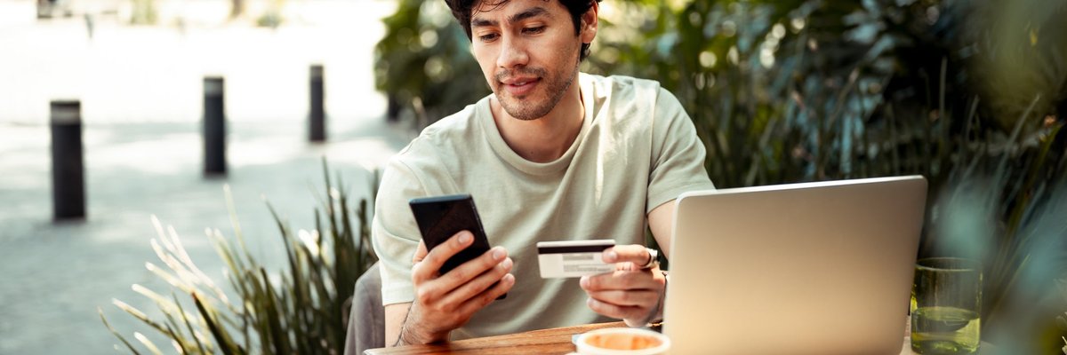 A person sitting at an outdoor cafe table holding a credit card and phone with open in front of them.
