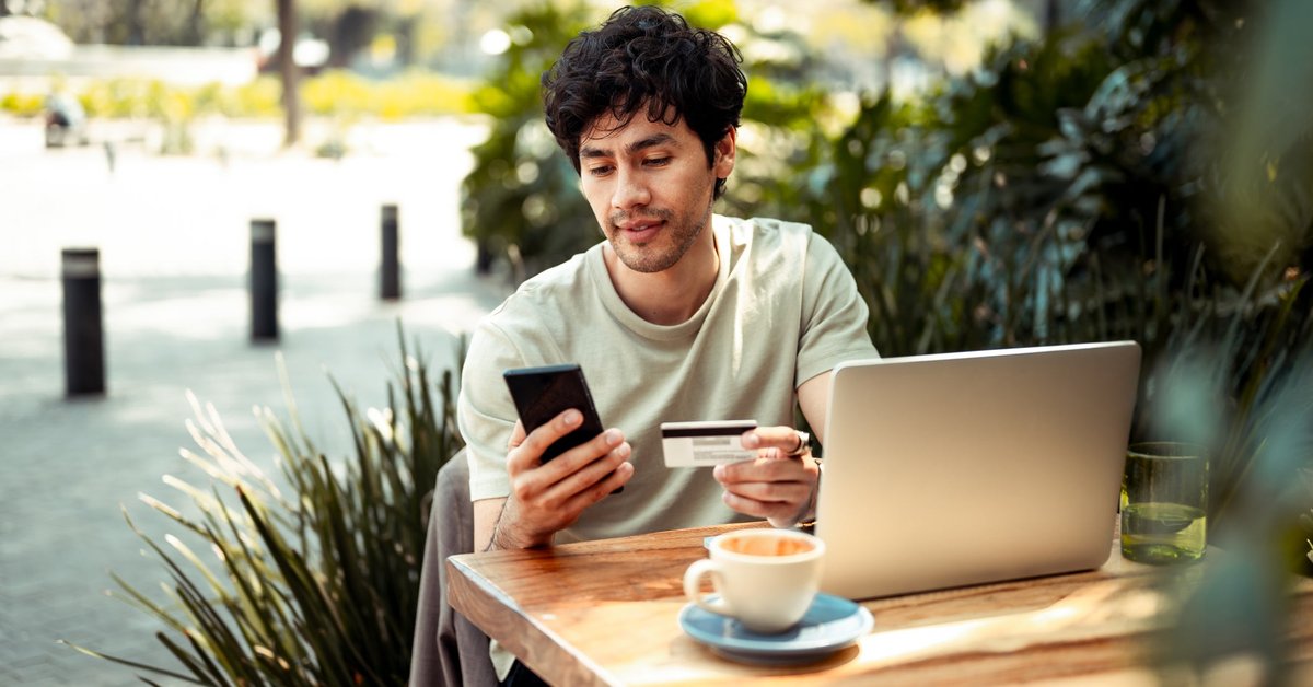 A person sitting at an outdoor cafe table holding a credit card and phone with open in front of them.