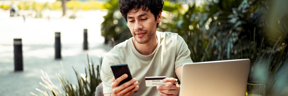 A person sitting at an outdoor cafe table holding a credit card and phone with open in front of them.