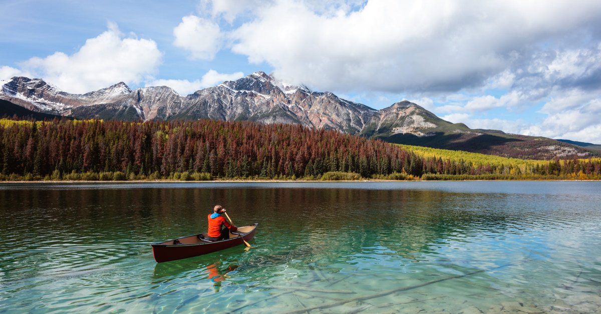 A person paddling a canoe across a clear blue lake surrounded by mountains in Canada.