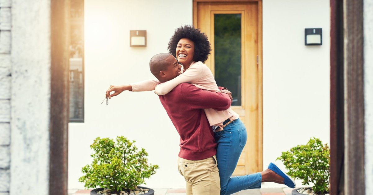 A person lifting another person in their arms who is holding keys to the new house behind them.