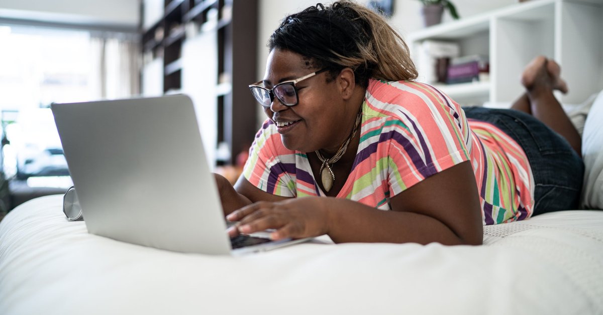 A smiling person lying on a bed while searching on a laptop.