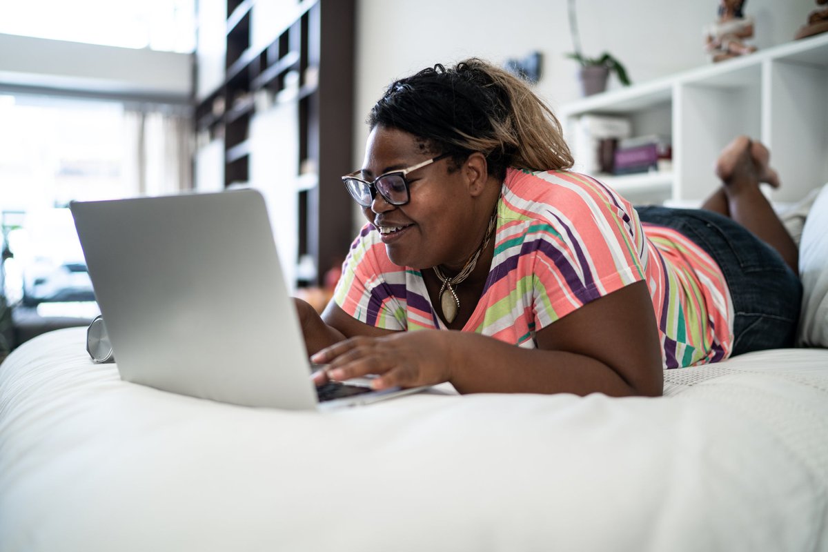 A smiling person lying on a bed while searching on a laptop.