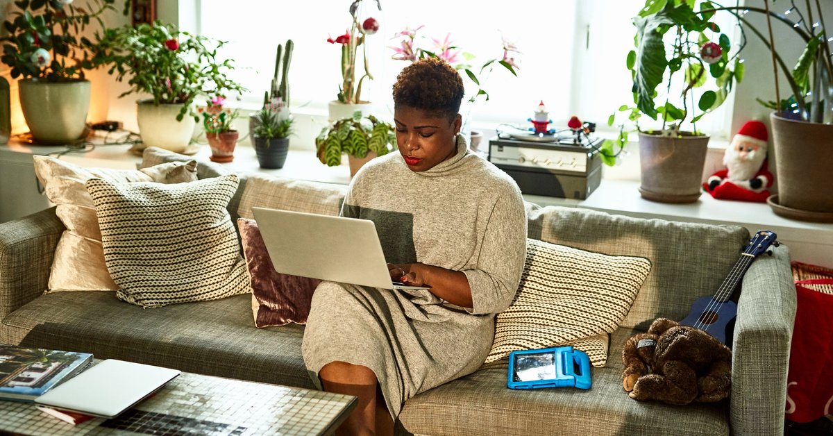 A person sitting on the couch in their living room surrounded by houseplants and typing on a laptop.