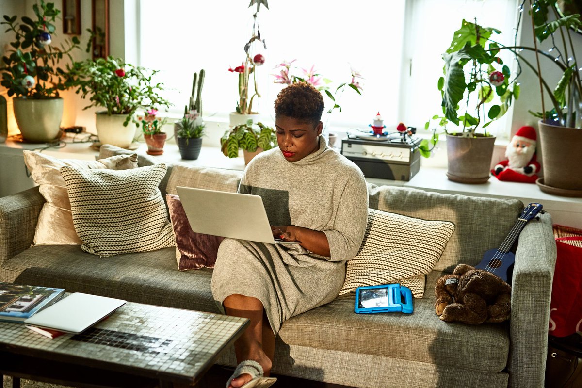 A person sitting on the couch in their living room surrounded by houseplants and typing on a laptop.