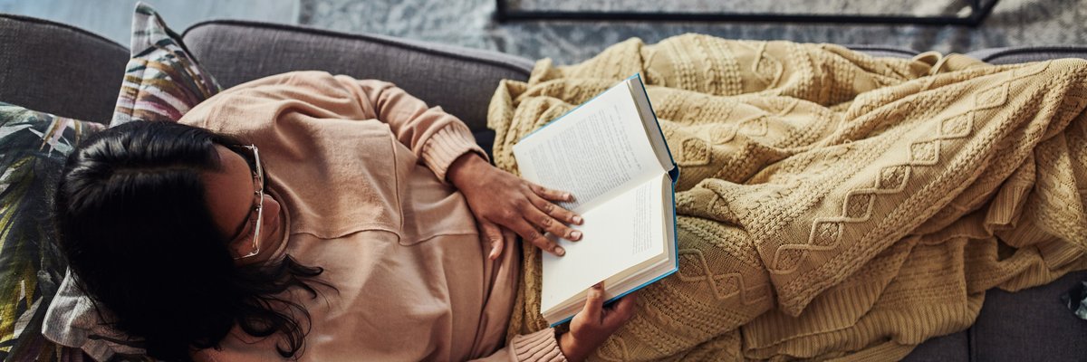 A person sitting on their couch under a blanket and reading a book.