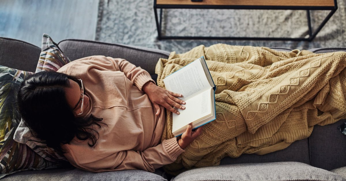 A person sitting on their couch under a blanket and reading a book.