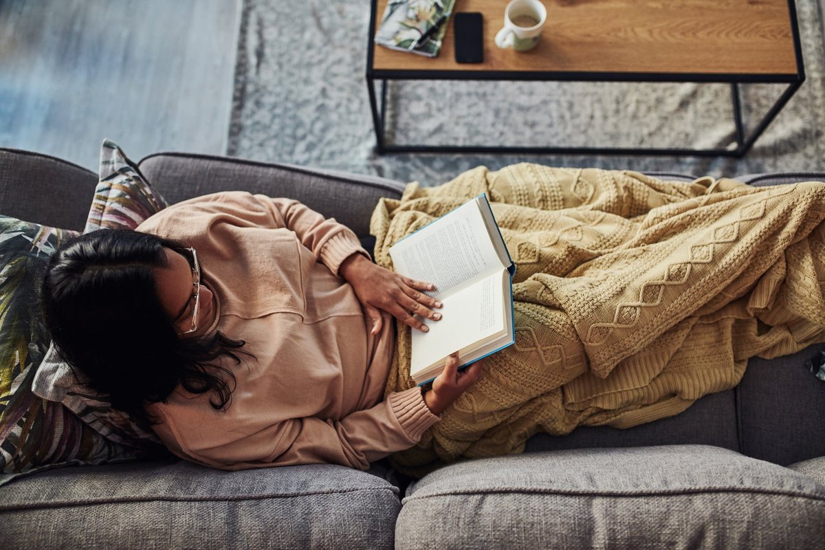 A person sitting on their couch under a blanket reading a book.