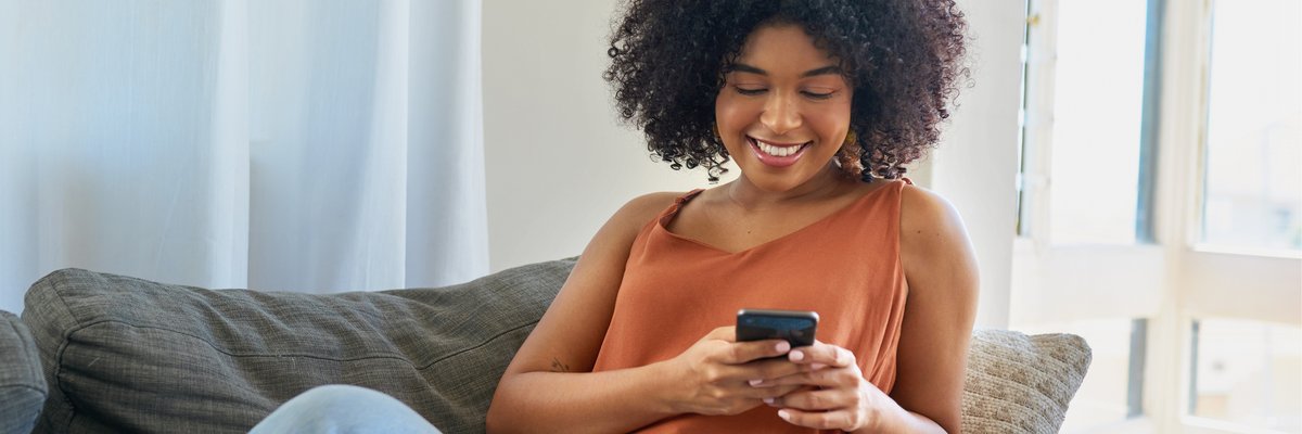 A smiling person sitting on their couch and looking at the phone in their hand.