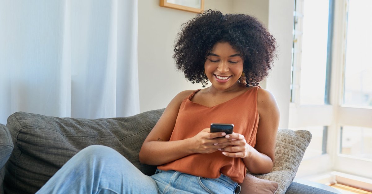 A smiling person sitting on their couch and looking at the phone in their hand.