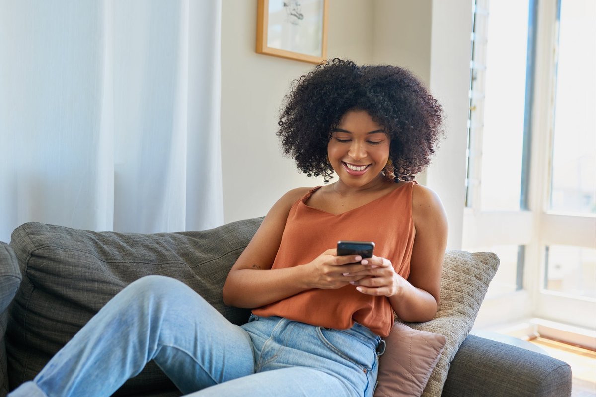 Een lachende persoon zittend op hun bank en kijkend naar de telefoon in hun hand.