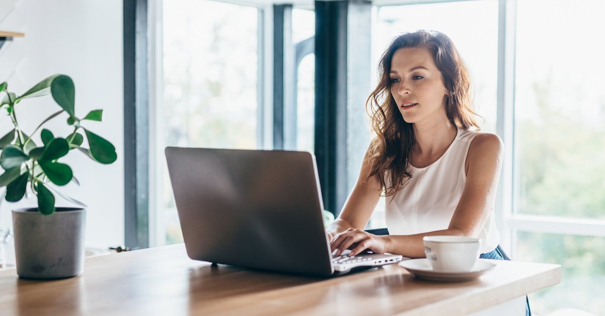 A person sitting at a sunny dining room table and typing on a laptop.