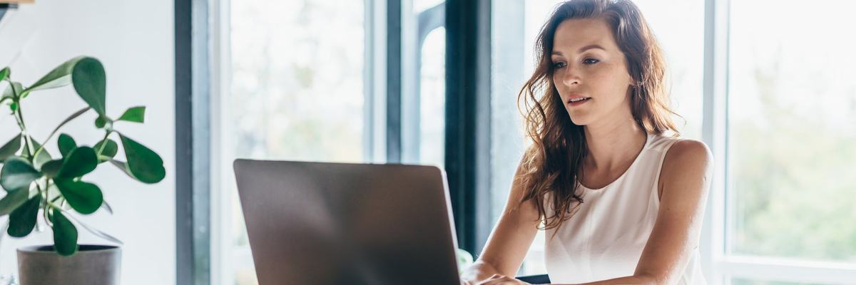 A person sitting at a sunny dining room table and typing on a laptop.
