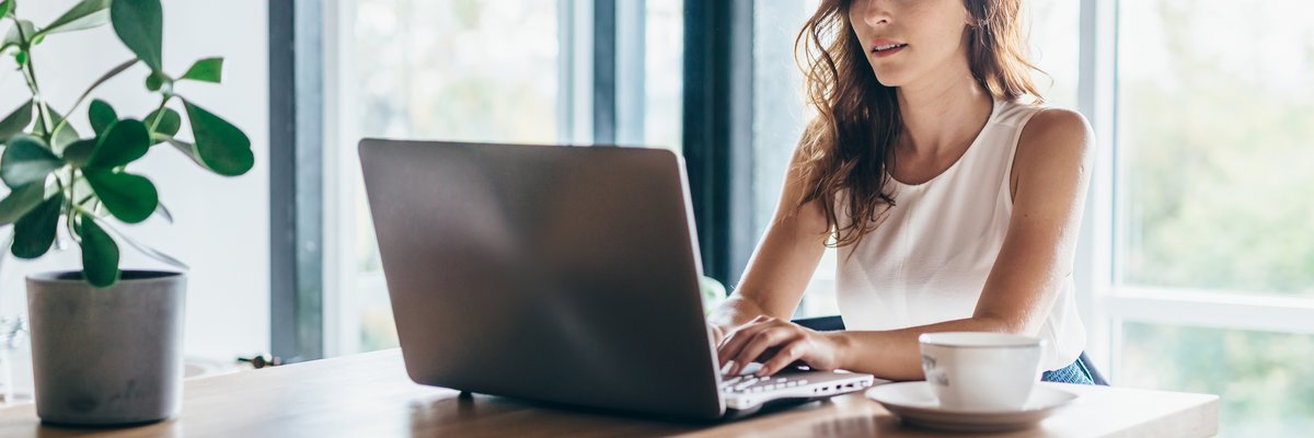 A person sitting at a sunny dining room table and typing on a laptop.
