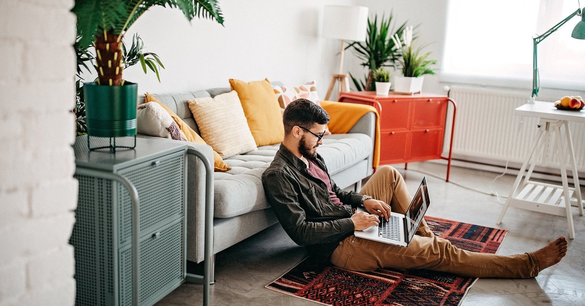 A person sitting on the floor leaning against a couch while typing on a laptop.