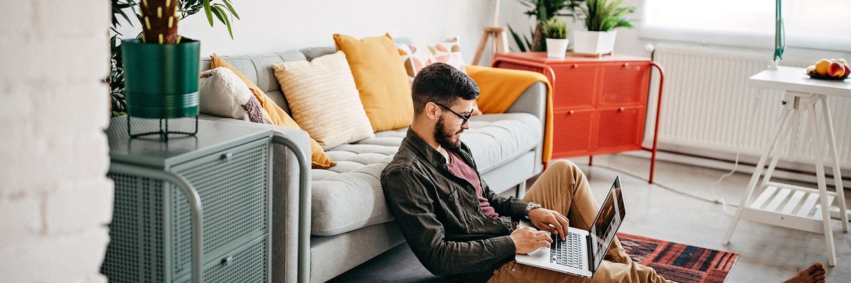 A person sitting on the floor leaning against a couch while typing on a laptop.