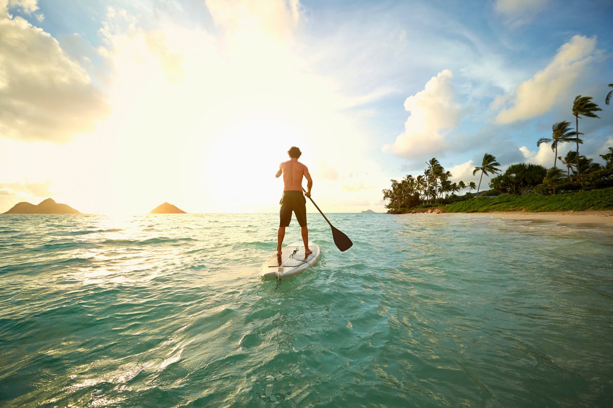 A person on a stand-up paddle board near a Hawaiian beach at sunset.
