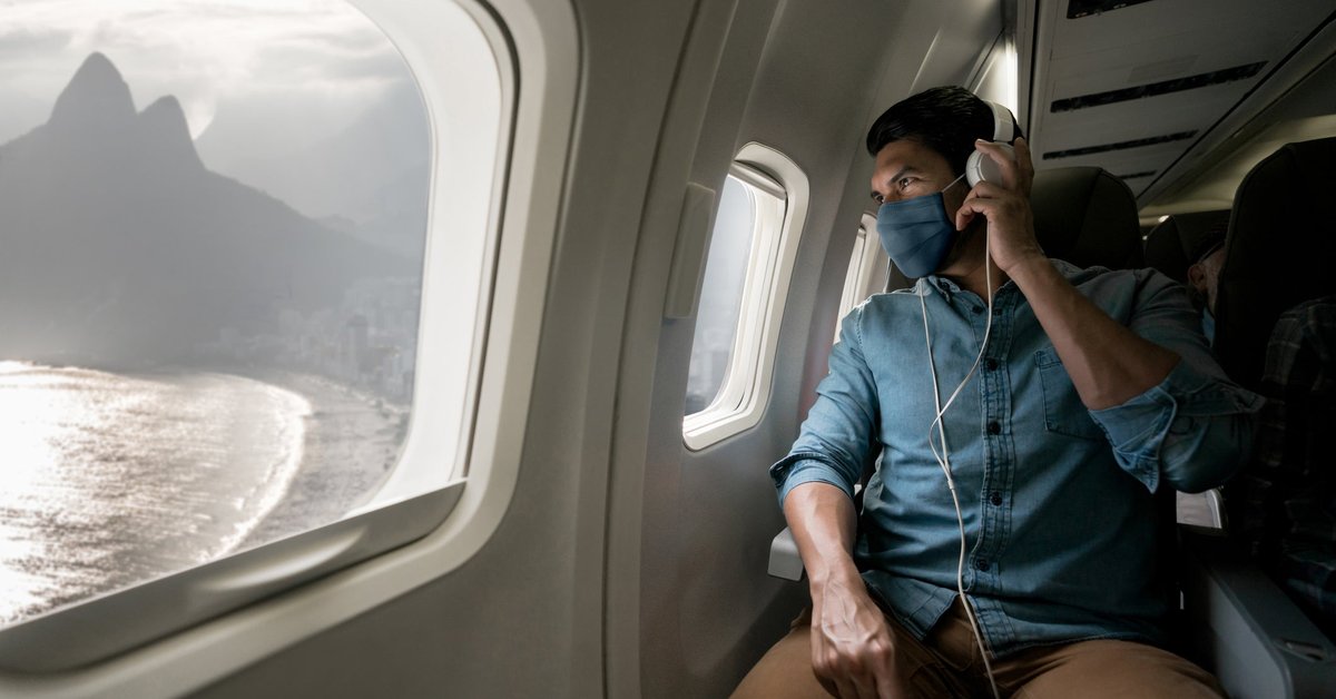 A person wearing a mask while sitting in a plane seat and looking out the window at a mountain next to the ocean.