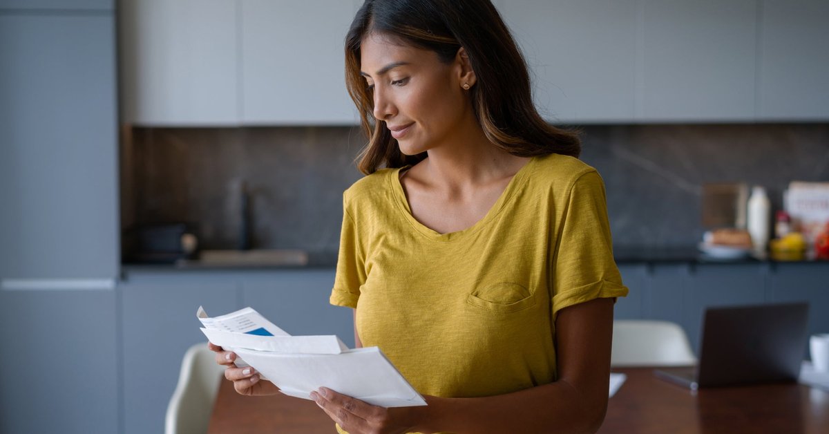 A smiling person opening their mail while standing in the kitchen.