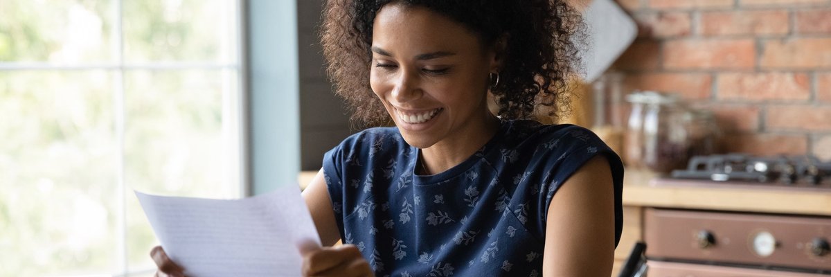 A person smiling while reading a piece of paper in their hand at their kitchen table.