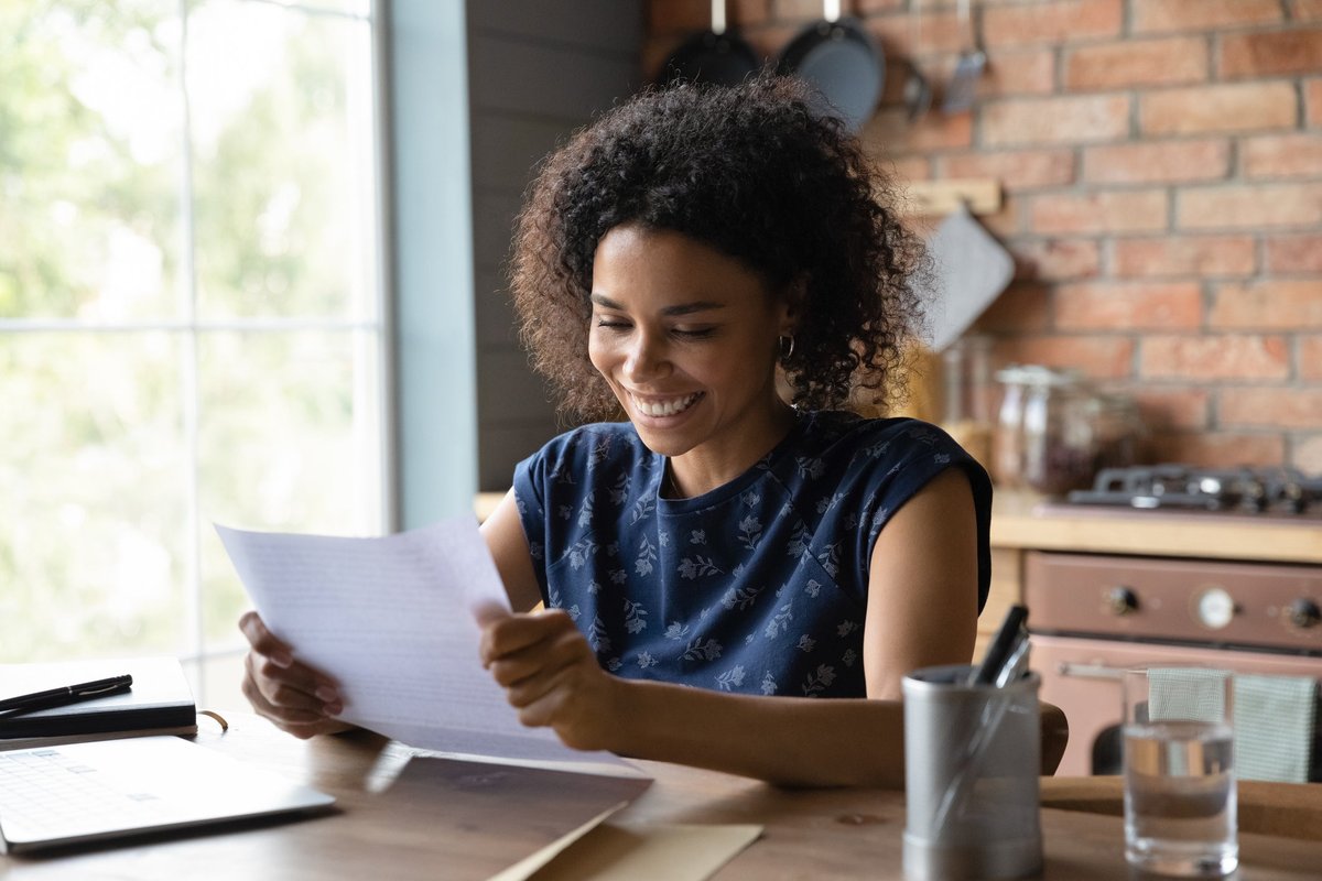 A person smiling while reading a piece of paper in their hand at their kitchen table.