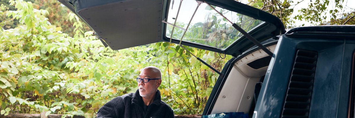 A person standing next to the open trunk door of a car and zipping up their jacket.