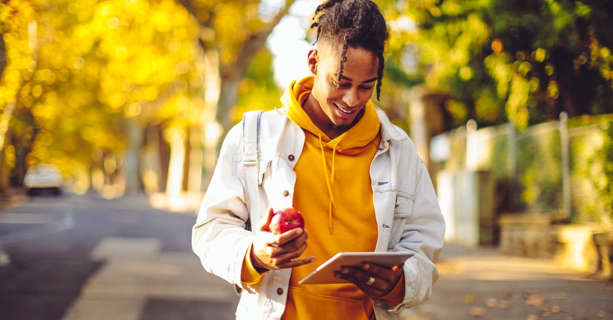 A person holding a tablet and eating an apple while walking through a park with fall trees.
