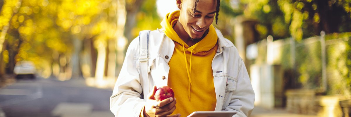A person holding a tablet and eating an apple while walking through a park with fall trees.