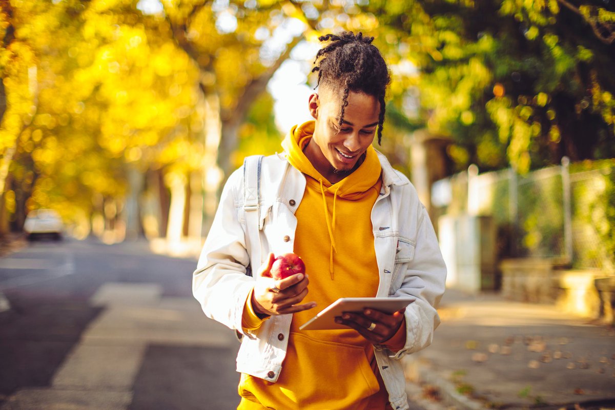 A person holding a tablet and eating an apple while walking through a park with fall trees.