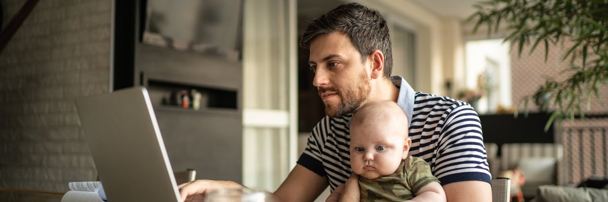 A person working on a laptop at a dining room table with a baby on their lap.