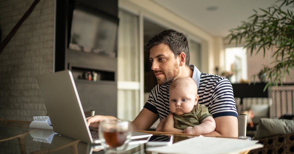 A person working on a laptop at a dining room table with a baby on their lap.
