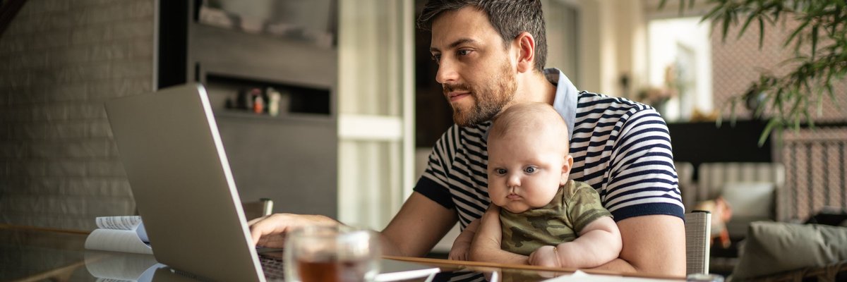A person working on a laptop at a dining room table with a baby on their lap.