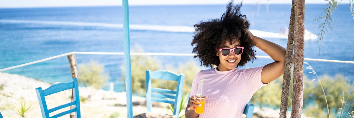 Person in sunglasses relaxing under umbrella at small table near the beach.