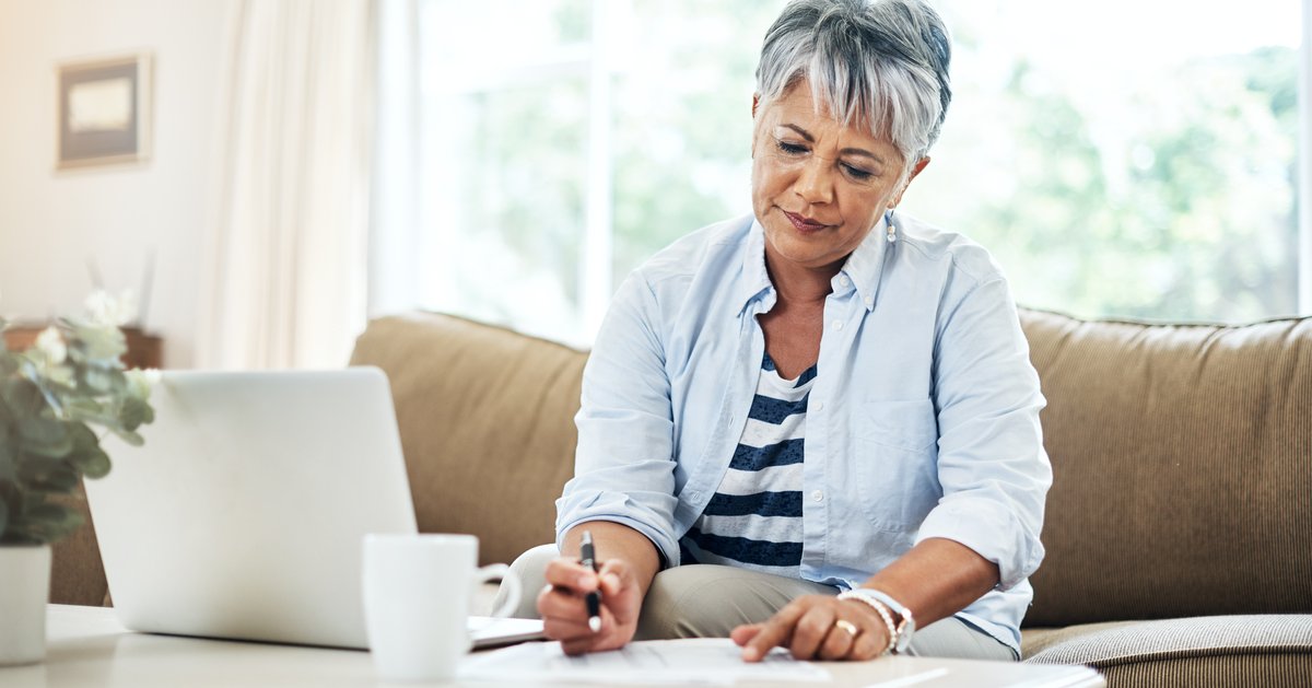 A person sitting on a couch and writing on a piece of paper next to a laptop open on a table.