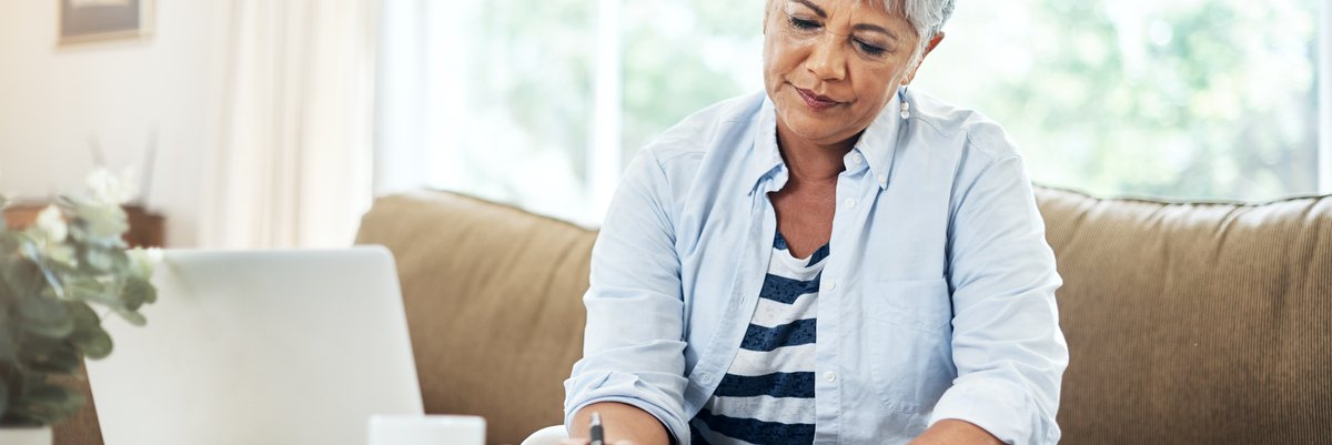 A person sitting on a couch and writing on a piece of paper next to a laptop open on a table.