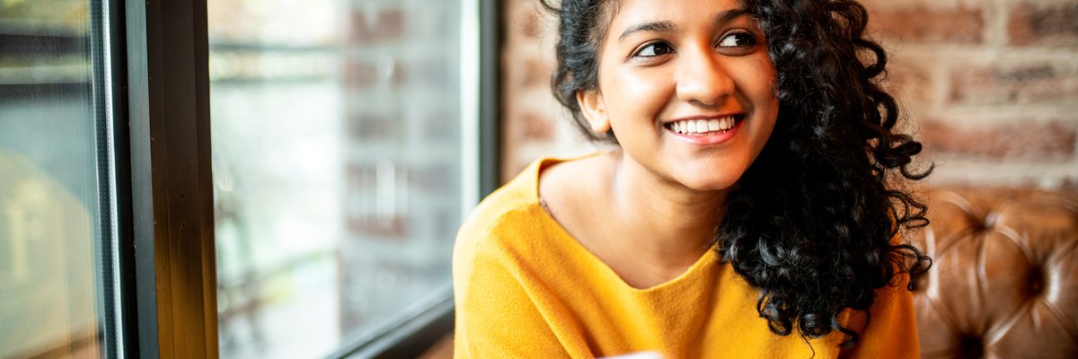A smiling person sitting on a couch near a window and holding a cell phone.