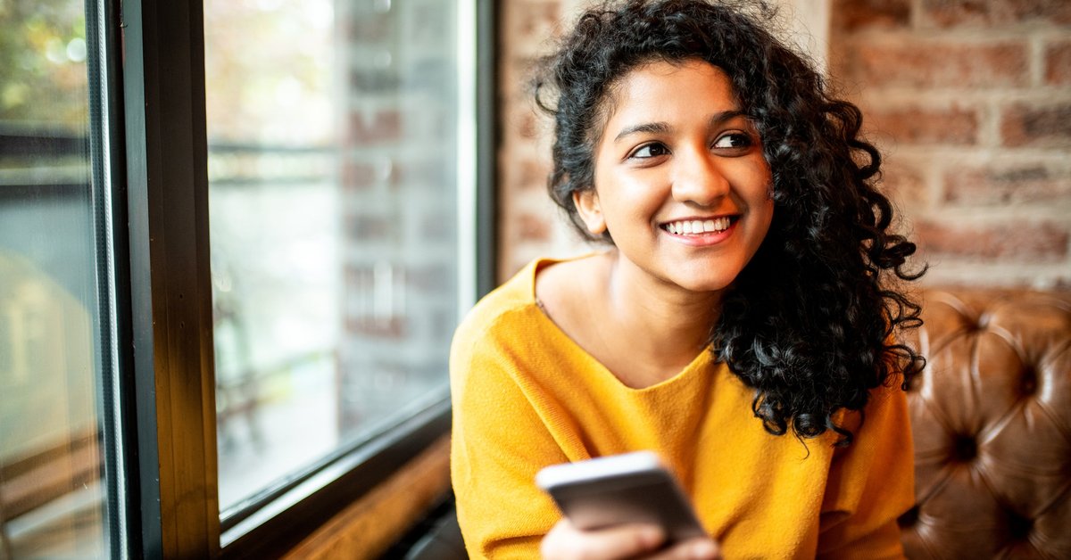 A smiling person sitting on a couch near a window and holding a cell phone.