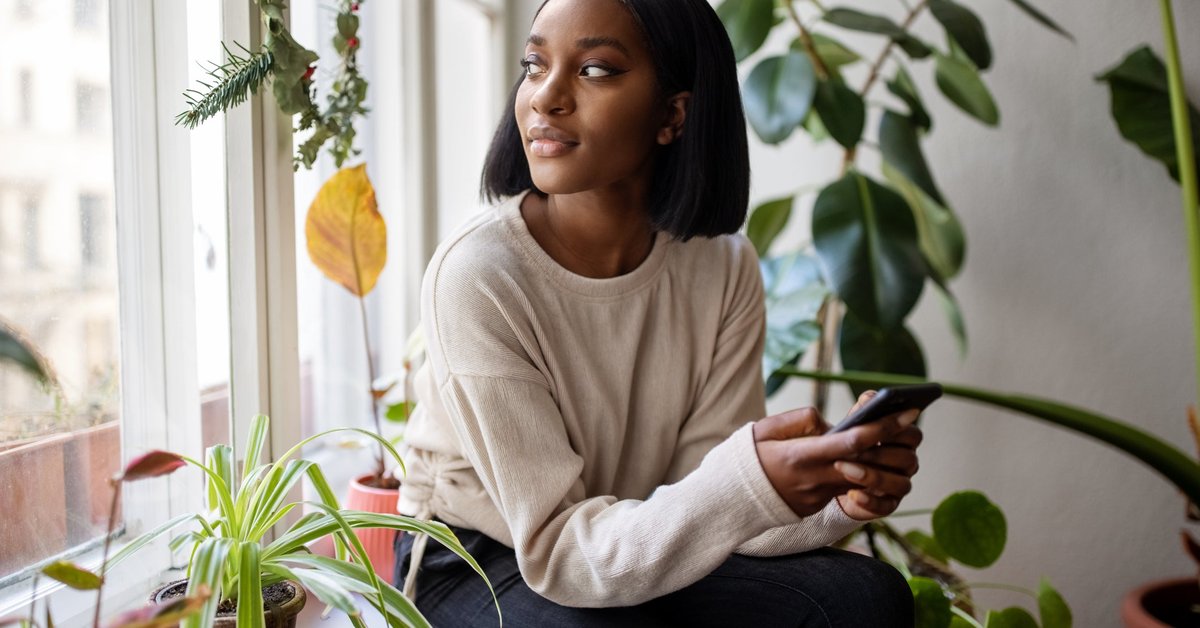A person holding a cell phone while looking out a window surrounded by house plants.