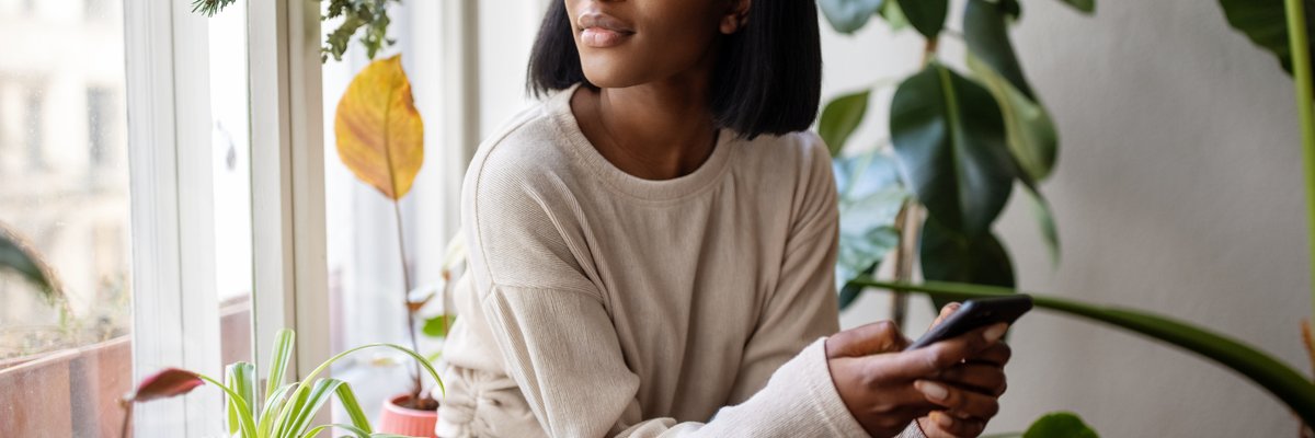 A person holding a cell phone while looking out a window surrounded by house plants.