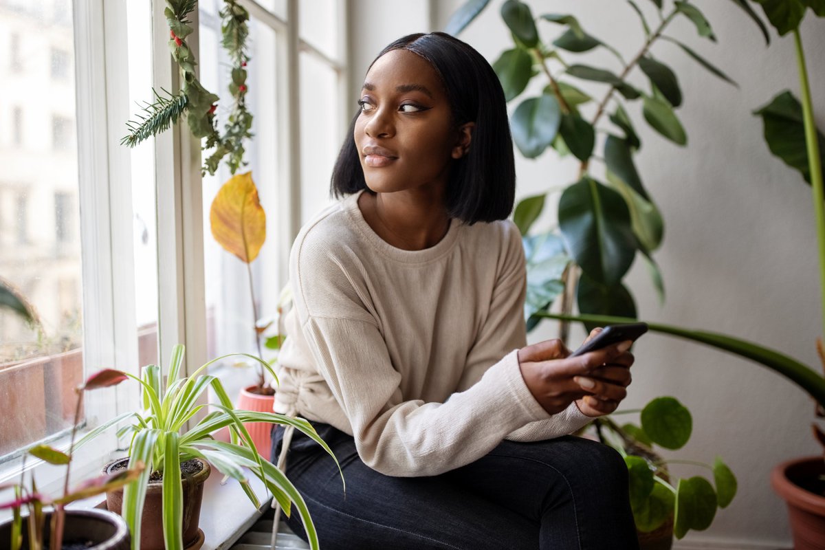 A person holding a cell phone while looking out a window surrounded by house plants.