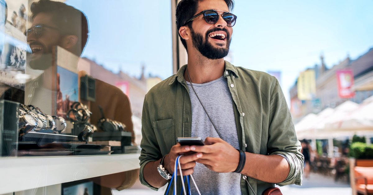 A smiling person holding a shopping bag and a phone while standing outside a store window filled with watches.
