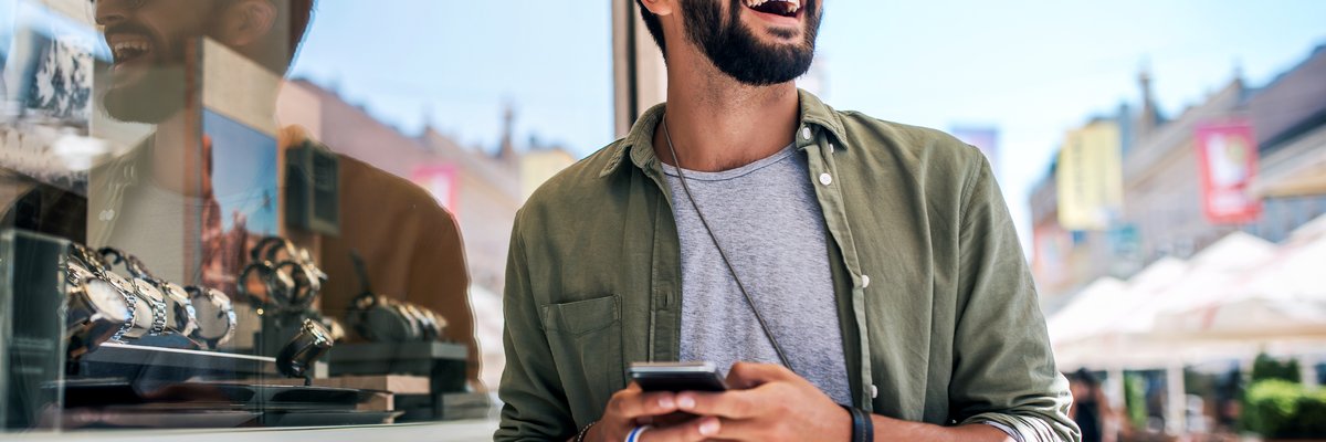 A smiling person holding a shopping bag and a phone while standing outside a store window filled with watches.