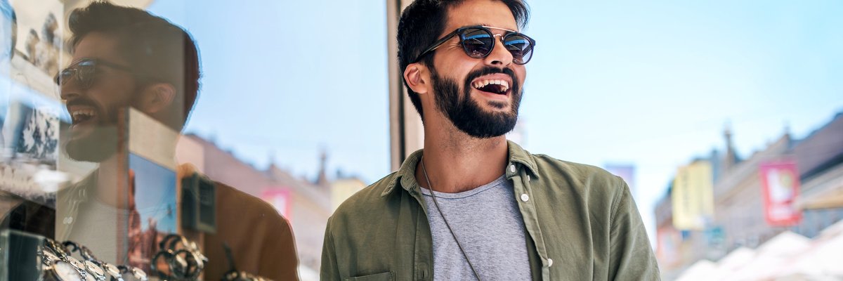 A smiling person holding a shopping bag and a phone while standing outside a store window filled with watches.