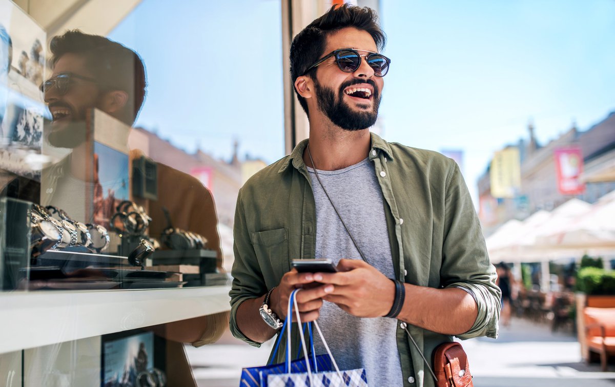A smiling person holding a shopping bag and a phone while standing outside a store window filled with watches.