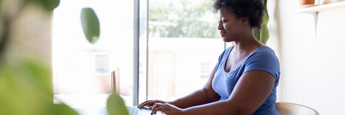 A person sitting at a desk in a sunny home office and typing on a laptop.