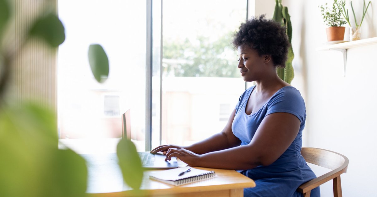 A person sitting at a desk in a sunny home office and typing on a laptop.